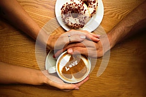 Romantic young couple sit in cafe,closeup hands holding a cup of coffee with heart on it and 2 donuts on wooden table background