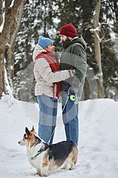 Romantic young couple in winter forest looking at each other and walking dog