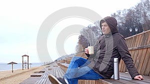 Romantic young man relaxing on the beach with , drinking hot tea or coffee from thermos. Calm and cozy evening.