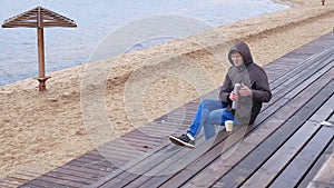 Romantic young man relaxing on the beach with , drinking hot tea or coffee from thermos. Calm and cozy evening.