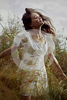 Romantic young woman in thick tall grass in field. Dreamy gentle woman in a white dress in nature. Natural beauty, enjoyment of