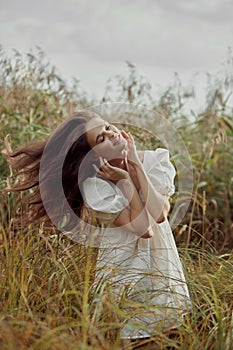 Romantic young woman in thick tall grass in field. Dreamy gentle woman in a white dress in nature. Natural beauty, enjoyment of