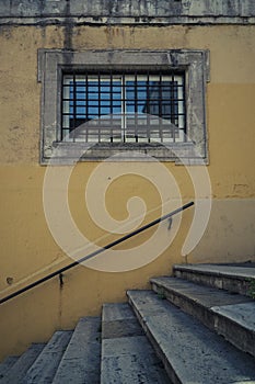 Stone window with metal bars, in a yellow wall with stairs underneath