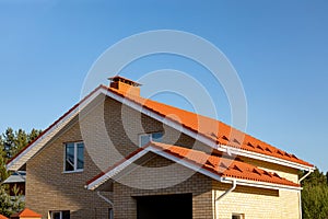 The roof of a brick house or cottage with slopes, tides, chimney against the blue sky. Roof made of red metal tiles for design on
