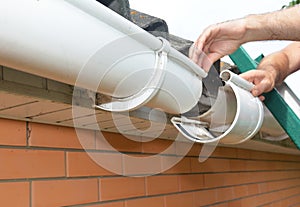 Roof gutter installation and repair. A man on a ladder is replacing a plastic rain gutter joint, bracket, connecting the gutters