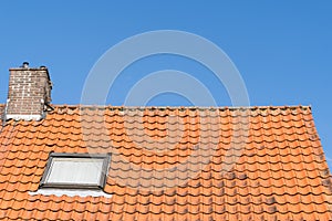 A Roof with red roof tiles, a window and chimney and a clear blue sky with some clouds on a sunny day