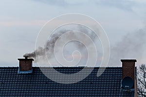 A roof with two chimneys on it. In one of the chimneys there is a pipe mounted on it were a lot of black polluting smoke is coming