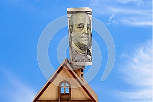 The roof of a wooden toy house with a dollar bill on a chimney against a blue sky