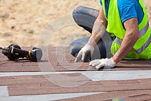 Roofer builder worker with nailgun installing Asphalt Shingles or Bitumen Tiles on a new house under construction.