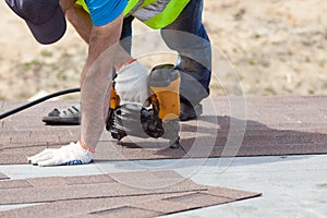 Roofer builder worker with nailgun installing Asphalt Shingles or Bitumen Tiles on a new house under construction.