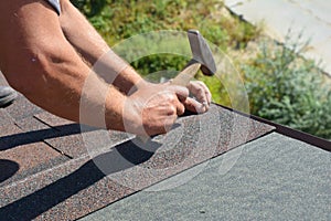 Roofer hands installing asphalt shingles on house construction roof corner with hammer and nails in motion.  Roofing Construction