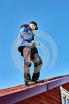 Roofer stands on roof with hammer in his hand against background of blue sky