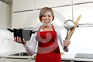 Rookie home cook woman in red apron at home kitchen holding cooking pan and rolling pin screaming desperate in stress