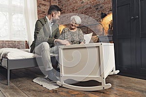 rookie middle-aged father and his grandmother quietly talking over a white crib where a little baby is napping