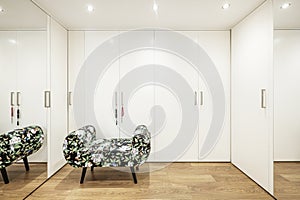 Room dedicated to a walk-in closet with smooth white wooden doors and large mirrors, wooden flooring and an upholstered armchair