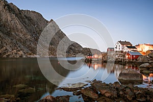 Rorbuer in Nusfjord in Lofoten islands in Norway during blue hour. Calm ocean and mountain peaks in the background.