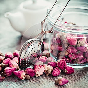 Rose buds tea, tea infuser, glass jar and teapot on background.