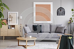 Rose gold painting and poster in bright loft interior with grey sofa near cabinet and wooden table. Real photo