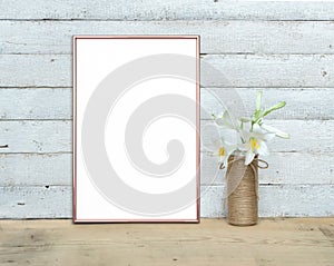 Rose Gold vertical A4 Frame mockup near a bouquet of lilies stands on a wooden table on a painted white wooden background. Rustic