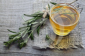 Rosemary herbal tea in a glass cup with fresh green rosemary herb on rustic wooden background.