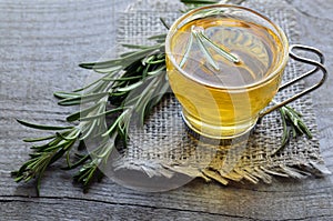 Rosemary herbal tea in a glass cup with fresh green rosemary herb on rustic wooden background.
