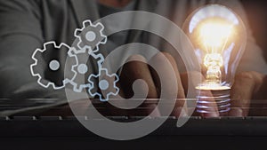 Rotating gears and a flashing light bulb against the background of a person working on a laptop in the dark with a glare