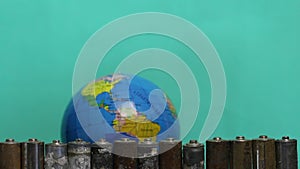 Rotating globe on a green background. In front of him is a row of spent batteries coated with corrosion. Recycling, World