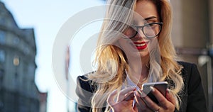 Rotation view of a gorgeous blonde woman in a formal wear and glasses standing outside the office and using her phone