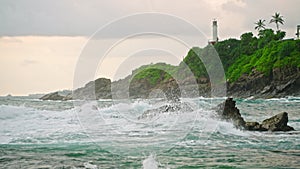 Rough waves crash against rocks near coastal lighthouse. Ocean storm, splash on cliff, beacon guides ships. Seascape