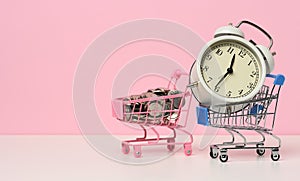 Round alarm clock in a miniature shopping cart with change on a white table. Concept time is money, waste of money and poverty