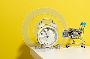 Round alarm clock, miniature shopping cart with coins on a white table. Concept time is money, waste of money and poverty