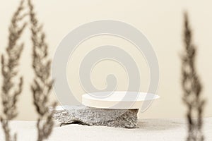 Round beige platform podium on grungy concrete stone on white beach sand with dry bent plant in foreground. Minimal