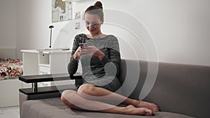 Round camera movement: smiling young woman sitting on the couch at home looking at phone and typing a message