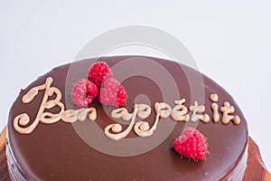 Round chocolate cake with raspberry decor and the inscription - Bon appetit. On a wooden stand isolated on a white background.