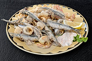 Round dish with mixed fried fish with sauce and lemon wedge on black s