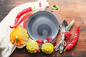 Round empty gray plate with space for text, yellow squash, red hot peppers, viburnum berries and cutlery on an old wooden surface