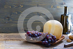 Round head of cheese Kostromskoy on textured dark wooden background on the square plate with grapes and defocused bottle