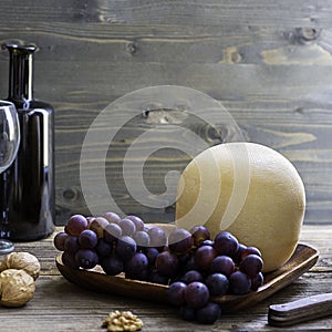 Round head of cheese Kostromskoy on textured dark wooden background on the square plate with grapes and defocused bottle