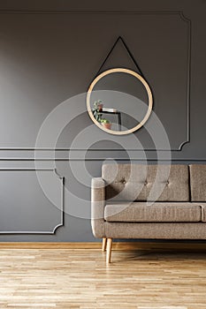 A round mirror above a simple, elegant brown sofa and a place for a side table in a fancy living room interior. Real photo.