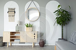 Round mirror hanging on the wall in real photo of grey living room interior with retro cupboard with lamp and books and fresh