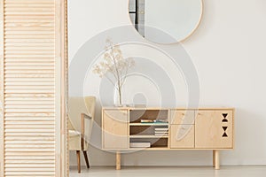 Round mirror on white wall above wooden cabinet in simple anteroom interior with armchair photo