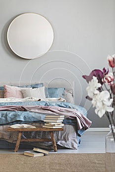 Round mirror in wooden frame on grey wall of elegant bedroom interior with comfortable bed with pastel, blue and pink bedding