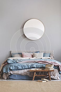 Round mirror in wooden frame on grey wall of elegant bedroom interior with comfortable bed with pastel, blue and pink bedding