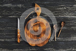 Round old cutting board with a wooden spoon and a knife on a dark rustic background. View from above.