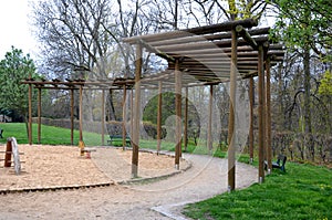 Round pergola, trellises made of wooden poles around the sandpit with play elements for children. around is a path and lawns. benc