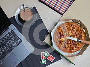 A round pizza, notebook, a cup of coffee are on a table.