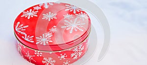 Round, red cookie and baked goods aluminum tin container decorated with white snowflake print pattern, resting on natural snow.