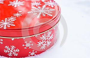 Round, red cookie and baked goods aluminum tin container decorated with white snowflake print pattern, resting on natural snow.