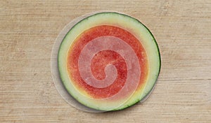 Round slice of pink seedless watermelon with a yellow and green rind, centered on a worn, pale brown cutting board