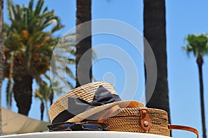 Round straw bag, straw hat and black sunglasses with palm trees and blue sky on the background